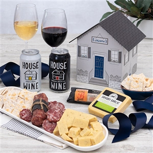 A gift basket that is shaped like a home with New Home printed on it filled with 2 cans of wine, cheese, crackers and soppressata.
